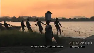 Local woman washing dishes pots on riverbank carrying them away on heads past local young.