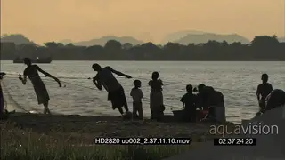 Local youn men pulling fishing nets out of river as the woman wash dishes nearby on.